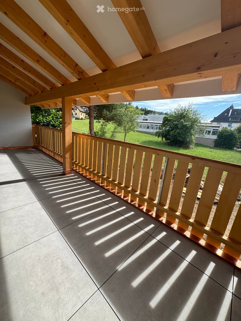 Balcony with wooden railing and tiled floor, overlooking a green lawn and houses in the distance.