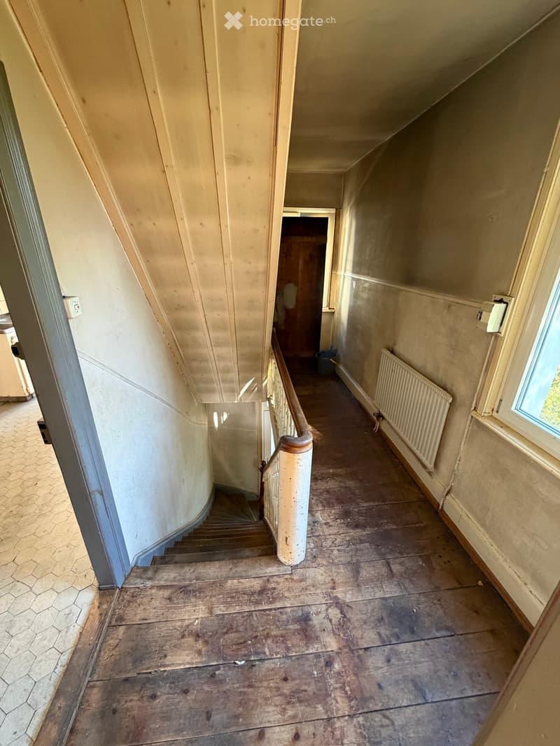 Empty hallway with wooden flooring, a staircase leading upwards, and a radiator on the right wall. The walls are painted in a light color, and there is a window at the end of the hallway.