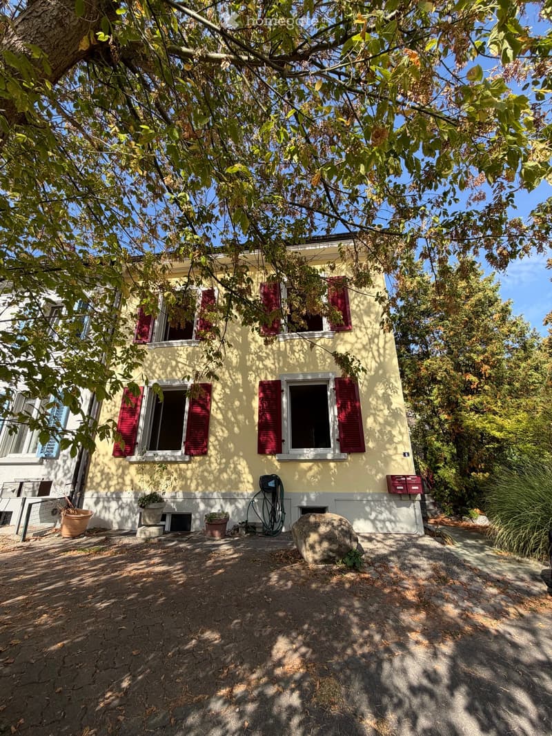 Two-story yellow house with red shutters, surrounded by trees and greenery, with a small garden area and potted plants near the entrance.