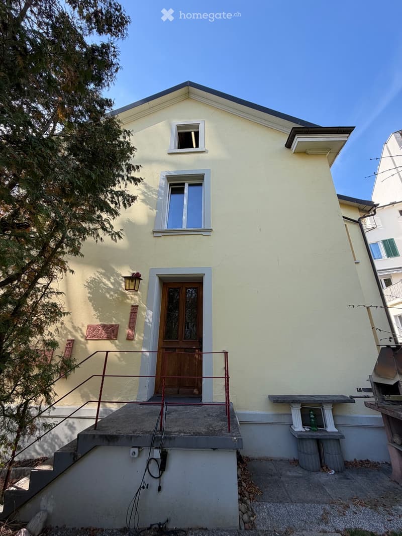 Two-story house with light yellow exterior, white-framed windows, wooden front door with decorative glass panels, small balcony with railing, and a stone bench on the side.