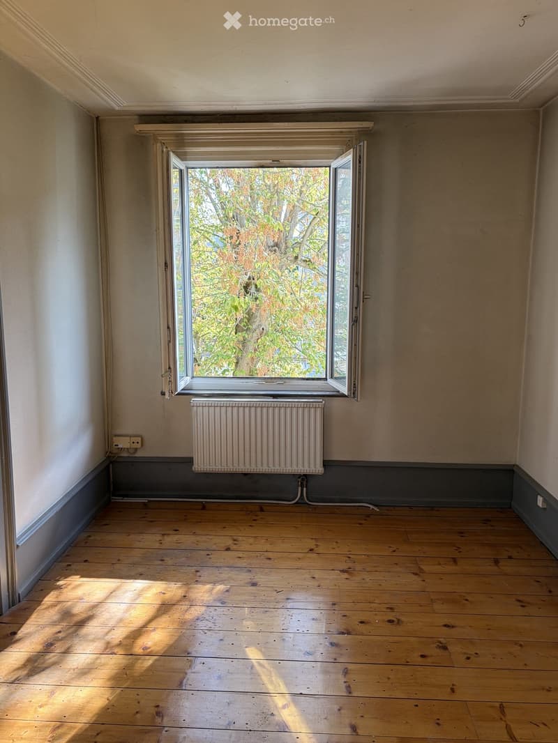 Empty room with wooden flooring, white walls, and a large window with open shutters allowing natural light to enter. A radiator is positioned below the window.