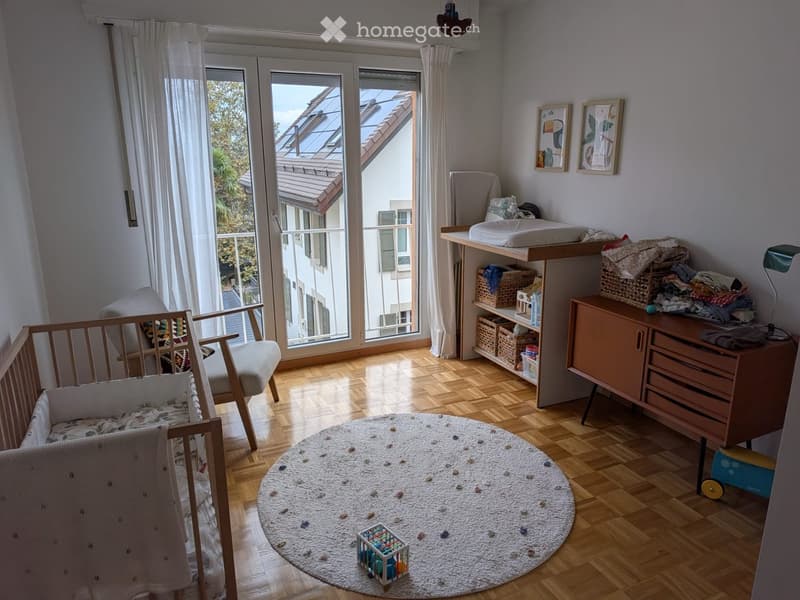 Brightly lit children's bedroom with wooden flooring, large windows, a crib, a chair, a cabinet, a table, and a round rug.