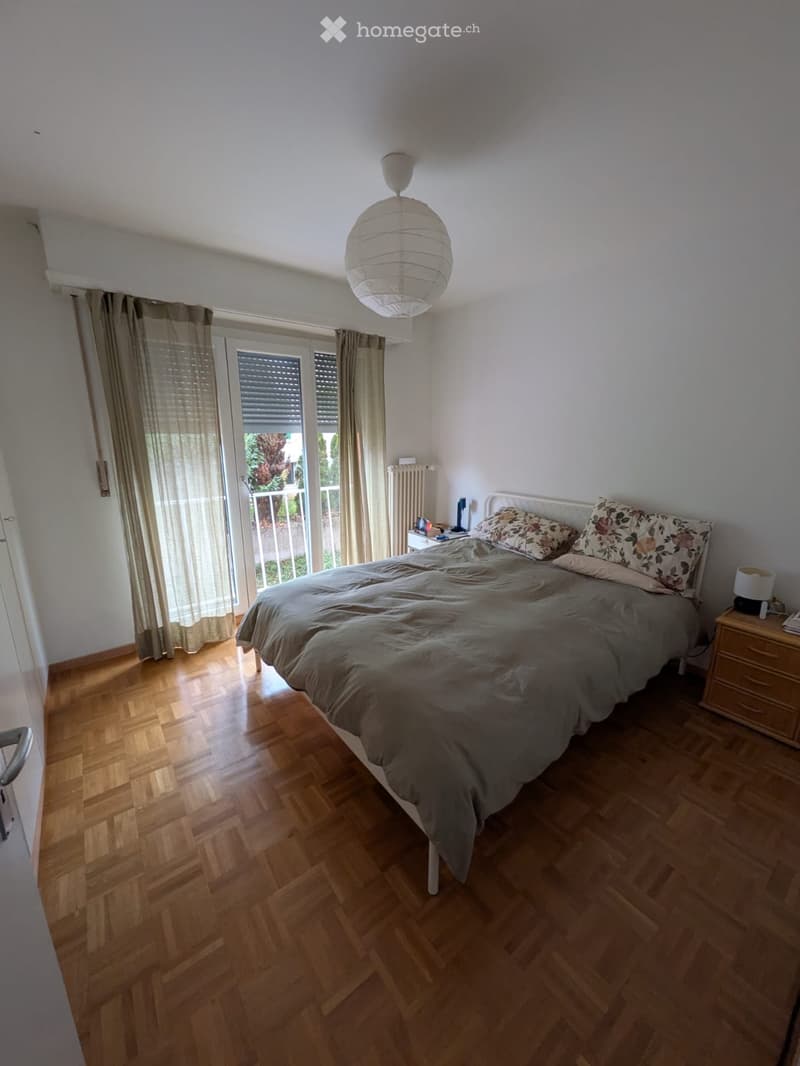 Cozy bedroom with wooden flooring, a large bed with patterned pillows and a fur throw, a nightstand with a lamp, and large windows with curtains leading to a balcony.