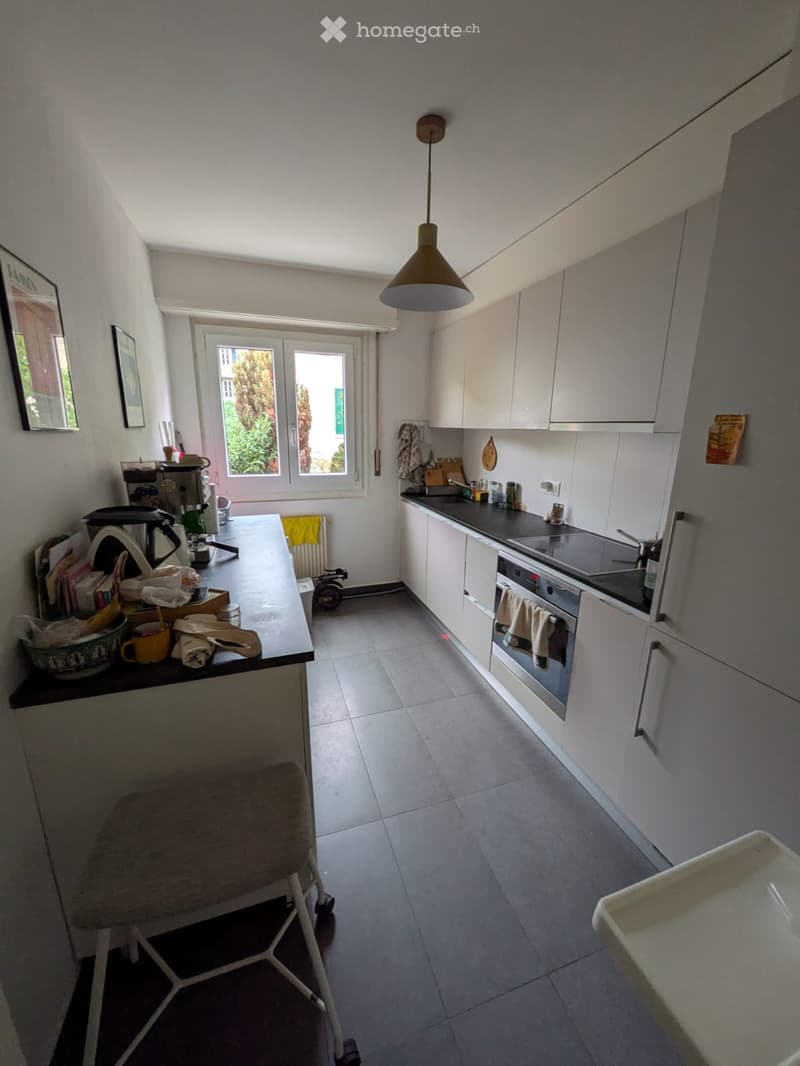 Modern kitchen with white cabinets, grey tiled floor, and a central island with a black countertop. Visible items include a stool, coffee maker, cups, and a framed picture on the wall.