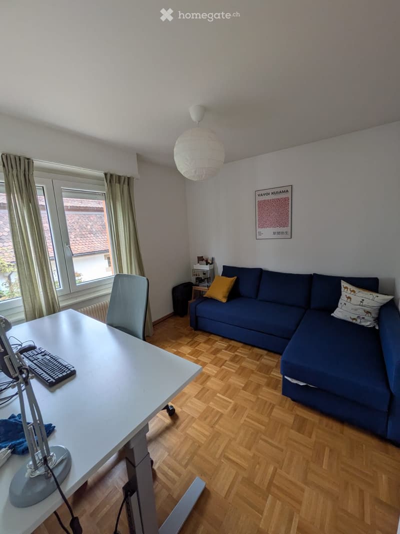 A modern living room with a blue sectional sofa, a white desk with a keyboard, a hanging paper lantern light fixture, and large windows with curtains.