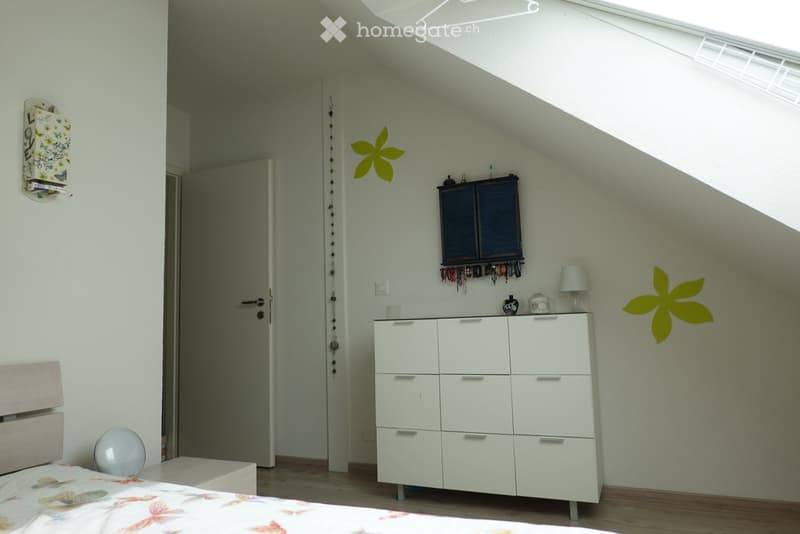Cozy attic bedroom with slanted ceilings, white walls, and wooden flooring. Features a bed with a floral duvet, a nightstand, a large white cabinet, a small lamp, and wall decorations including a yellow flower sticker and a mirror.