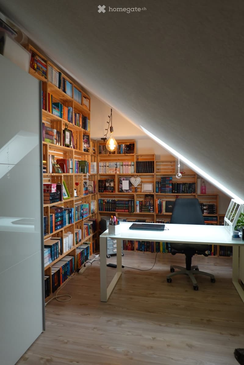 A cozy attic study with wooden bookshelves filled with books, a white desk with a laptop, a black office chair, and a hanging light bulb.