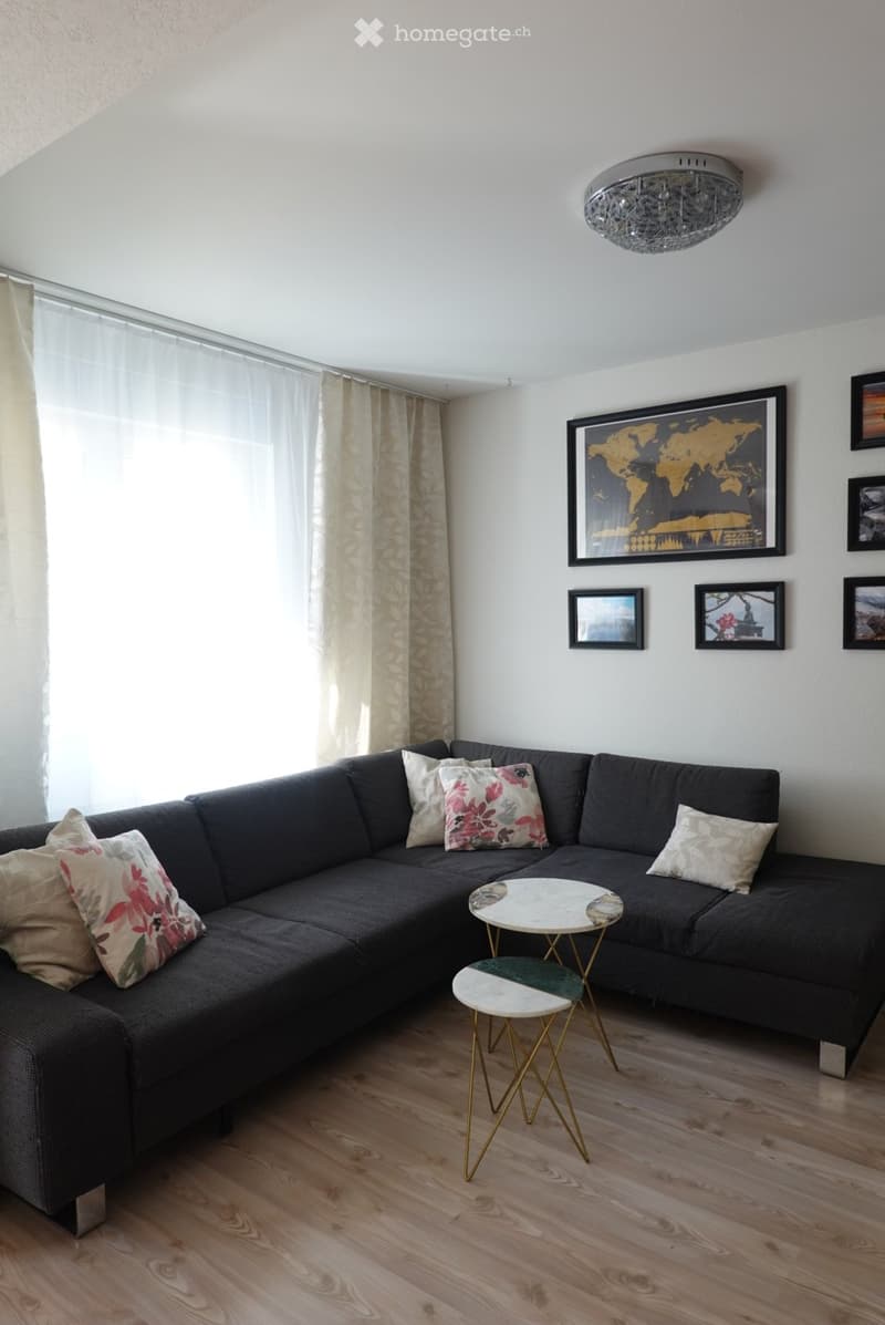 Modern living room with a dark grey sectional sofa, decorative pillows, a small round coffee table, and a matching stool. The room features light wooden flooring, large windows with sheer curtains, and several framed pictures on the wall.