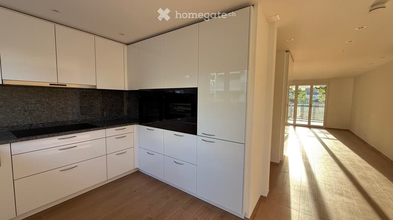 Modern kitchen with white cabinets, granite countertop, and stainless steel appliances, leading to a sunlit living space with wooden flooring.