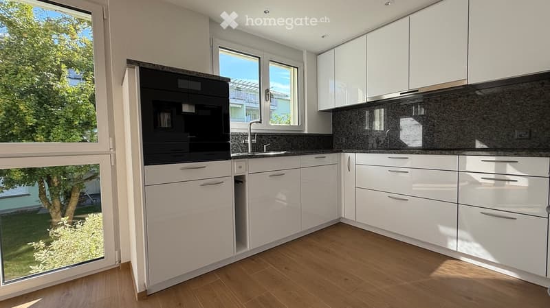 Modern kitchen with white cabinets, granite countertops, and a built-in oven. Large windows provide natural light and a view of the garden.