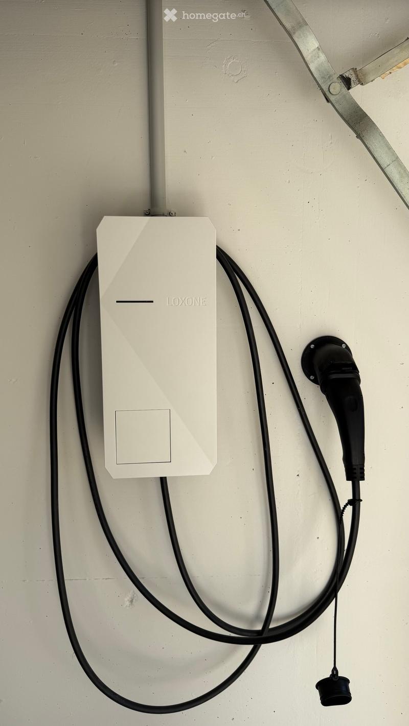 A white Loxone electric vehicle charging station mounted on a wall with cables connected.