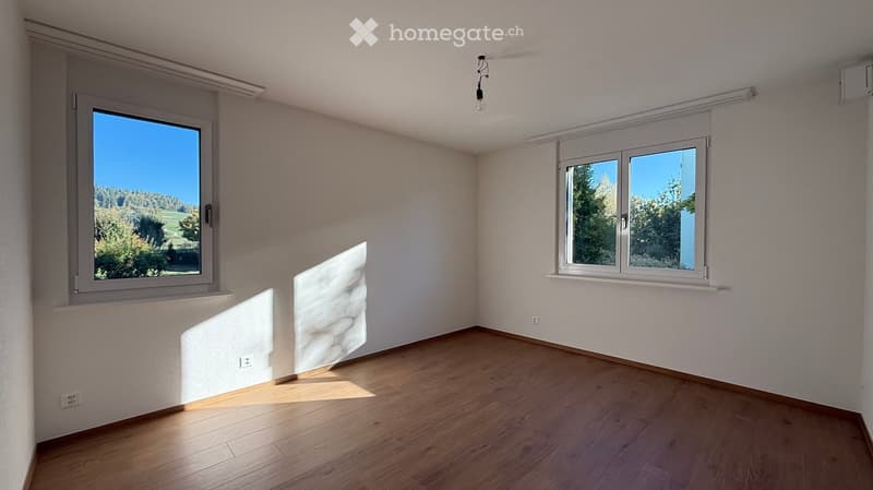 Empty room with wooden flooring, white walls, and large windows offering a view of greenery outside.