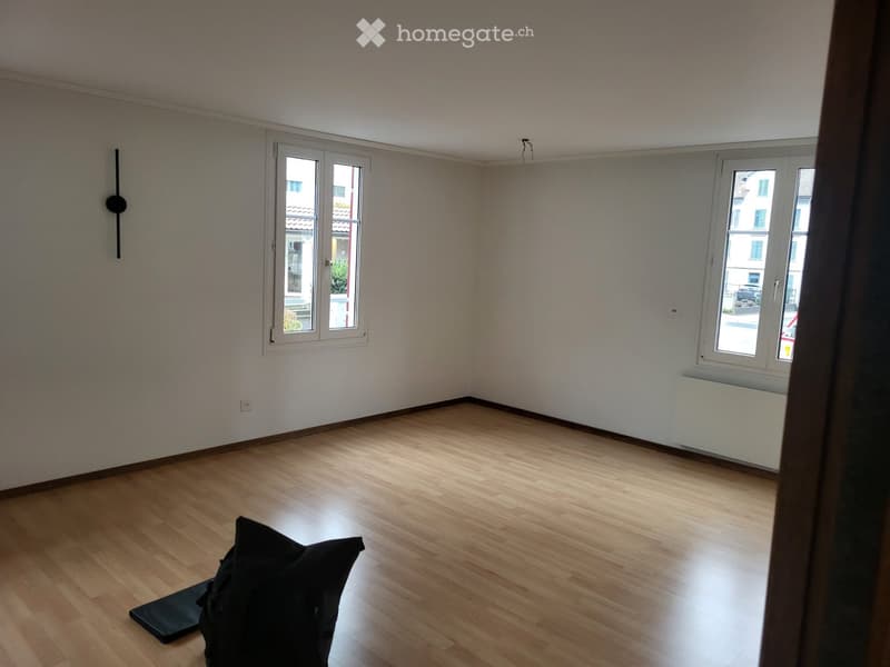 Empty room with wooden flooring, white walls, and two windows offering a view of the street.