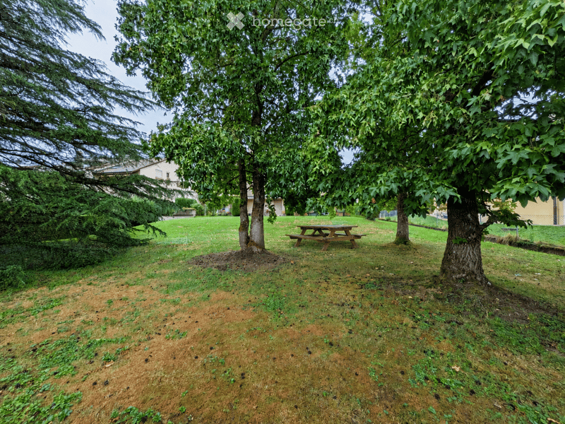 A grassy outdoor area with several large trees, a wooden picnic table, and a building visible in the background.