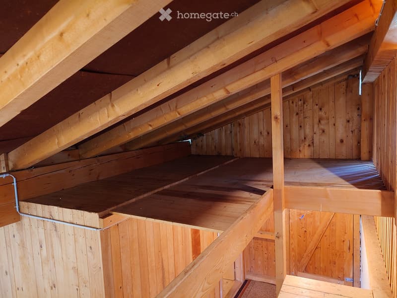 An attic space with exposed wooden beams and unfinished wooden walls, featuring built-in storage shelves and a staircase leading up.