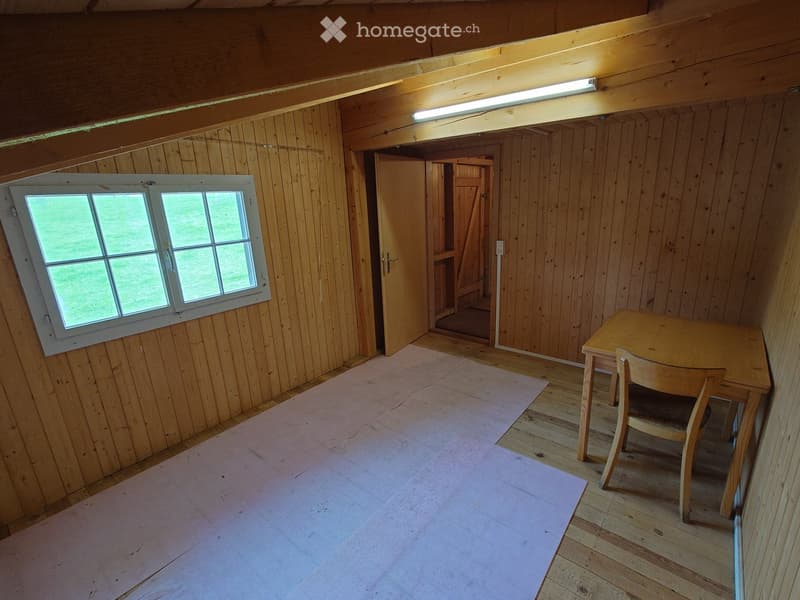 A small wooden room with a slanted ceiling, a window, a table, and a chair. The floor is wooden, and there is a pink protective sheet on part of the floor.