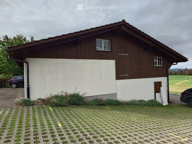 Two-story house with a combination of wooden and white walls, multiple windows, and a tiled roof. The house is surrounded by a paved area with grass patches and has two parked cars nearby.