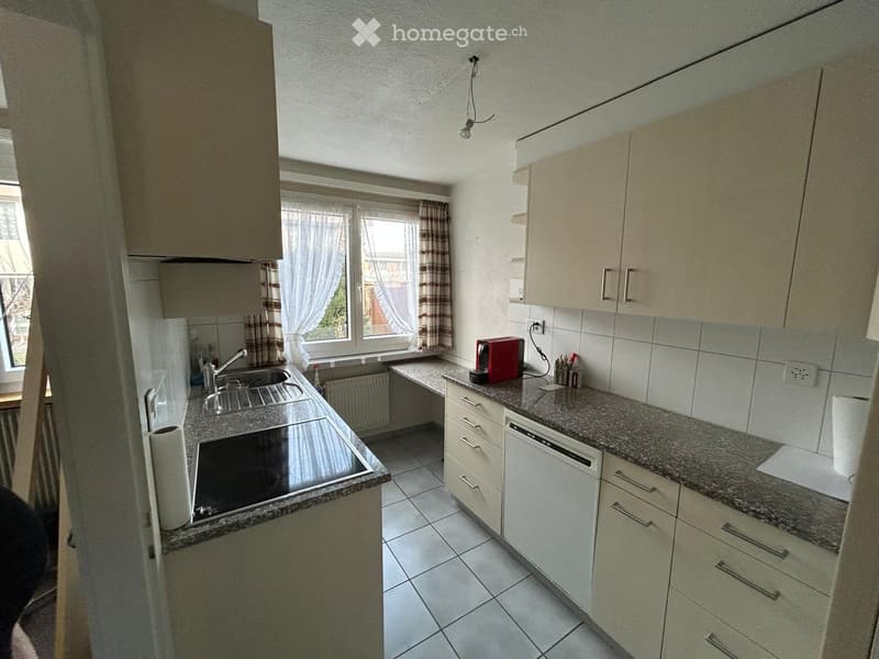 Modern kitchen with light-colored cabinets, granite countertops, and stainless steel appliances including a sink, stove, and dishwasher. A window with curtains allows natural light to enter.