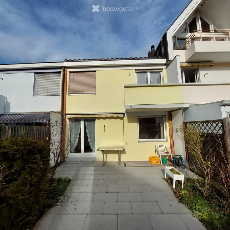 Garden area with tiled patio, small table, chairs, and storage items near a house with yellow and white exterior.