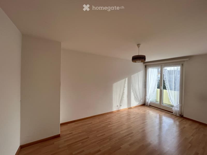 Empty room with wooden flooring, white walls, and large windows with white curtains. A hanging light fixture is visible.