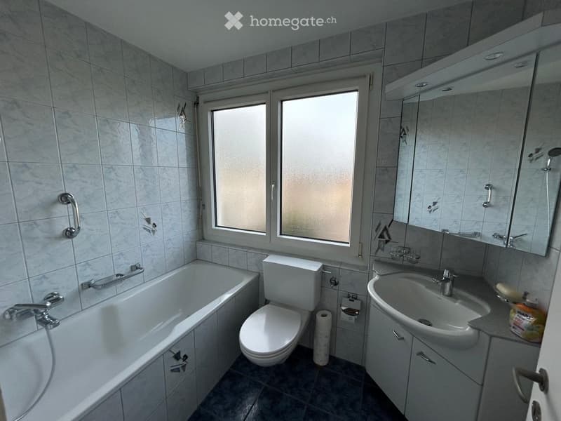 Bathroom with a bathtub, toilet, sink, mirror, and cabinet. The room has tiled walls and a window with frosted glass.