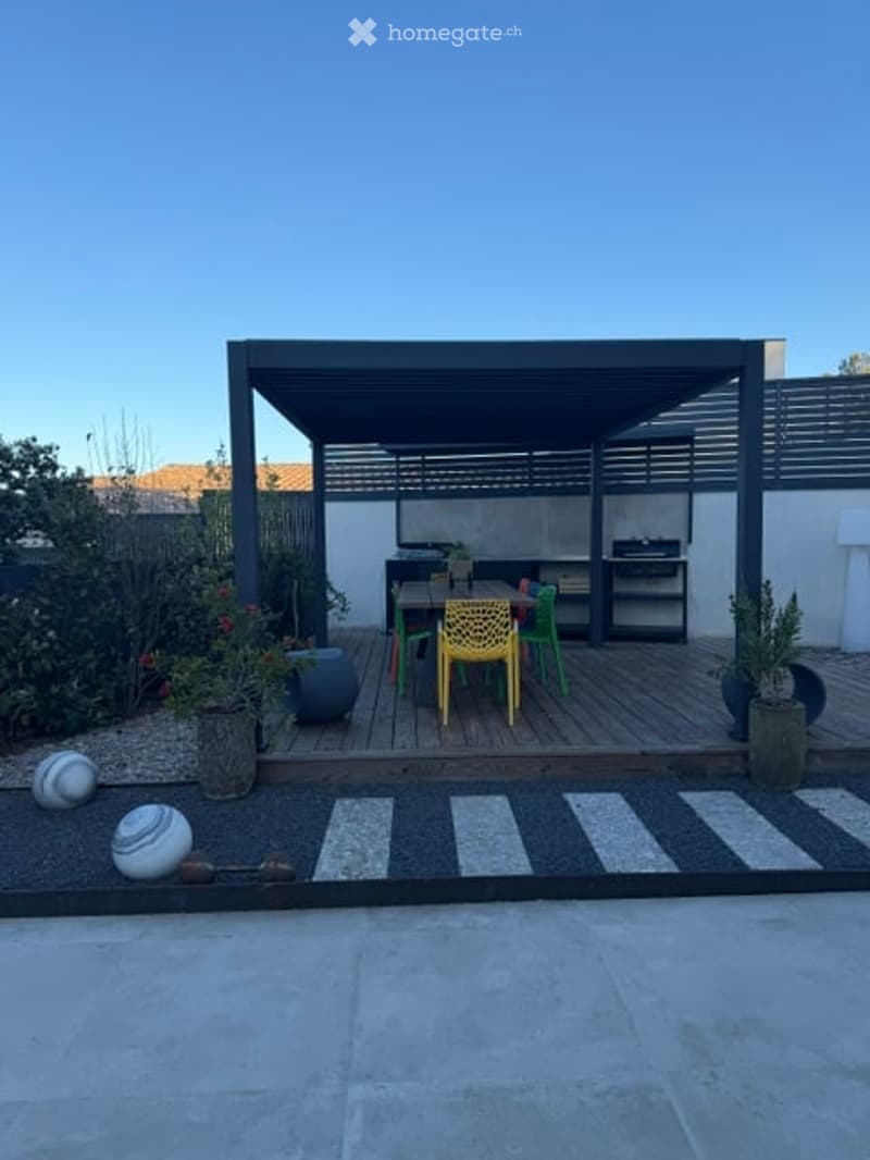 A modern outdoor patio with a wooden deck, a black pergola, and colorful chairs arranged around a small table. Potted plants and decorative balls are placed around the area.