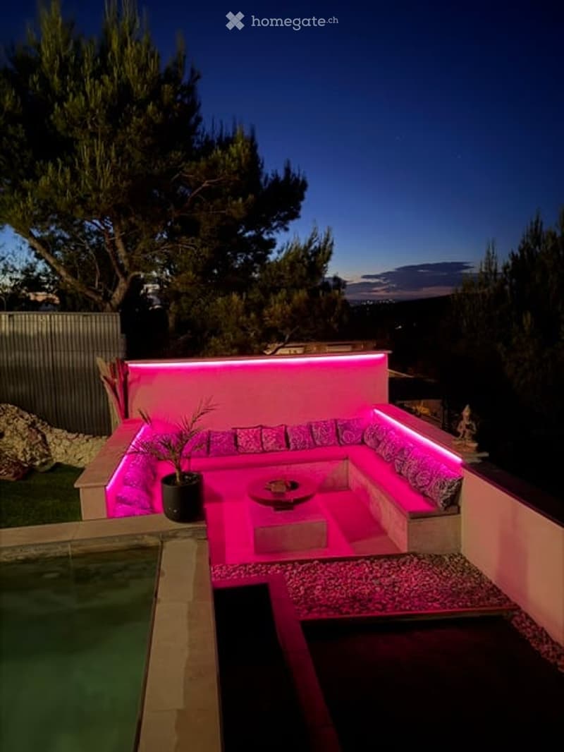 Modern outdoor seating area with built-in sofa illuminated by pink neon lighting, a small coffee table, and a potted plant, adjacent to a swimming pool.