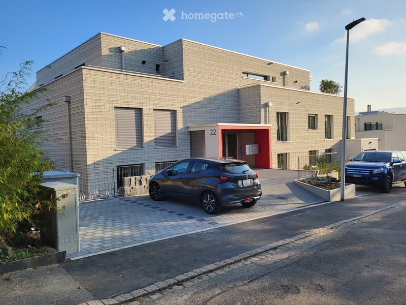Modern two-story building with brick facade, large windows, and a red entrance area. Two cars are parked in front, and there is a small landscaped area with plants and a streetlight.