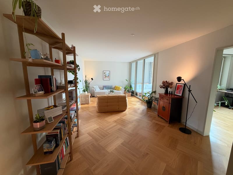 Bright living room with wooden herringbone flooring, large window, bookshelf, sofa, ottoman, and decorative items.