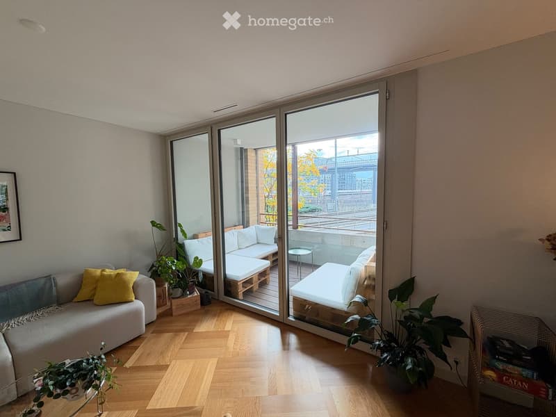 Bright living room with wooden flooring, large sliding glass doors opening to a balcony, grey sofa with yellow cushions, potted plants, and a small table on the balcony.