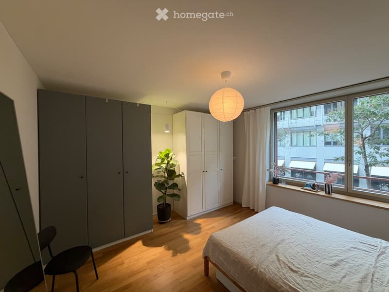 Modern bedroom with a wooden floor, large window with white curtains, a bed with white bedding, a black chair, a grey wardrobe, and a potted plant near the window.