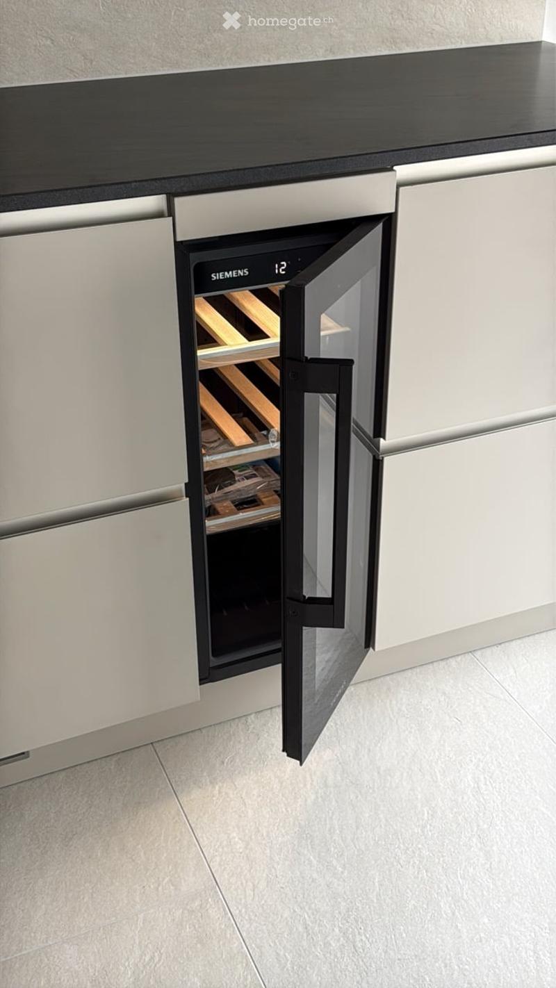 A modern kitchen with sleek white cabinets and a black countertop, featuring an open Siemens wine cooler with wooden racks inside.