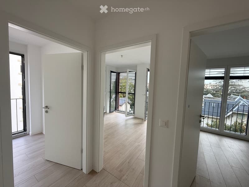 Empty hallway with light wooden flooring, white walls, and multiple doors leading to different rooms and a balcony area.