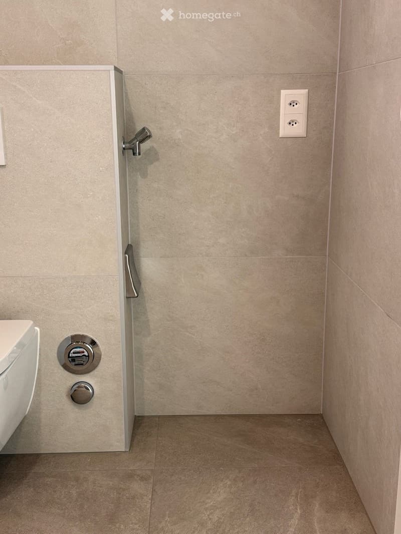Modern bathroom with beige tiled walls and floor, featuring a toilet, a chrome faucet, and a power outlet on the wall.