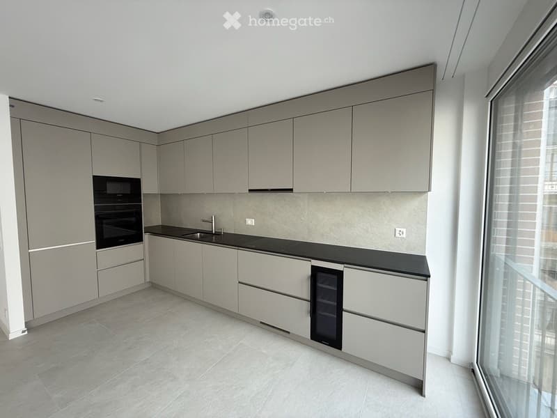 Modern kitchen with light grey cabinets, black countertops, and built-in appliances including an oven and refrigerator. Features a large window with blinds and a sleek, minimalist design.