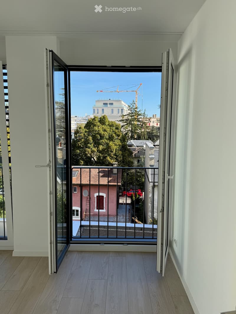 Balcony with wooden flooring, glass doors, and a metal railing, overlooking a cityscape with trees and buildings.