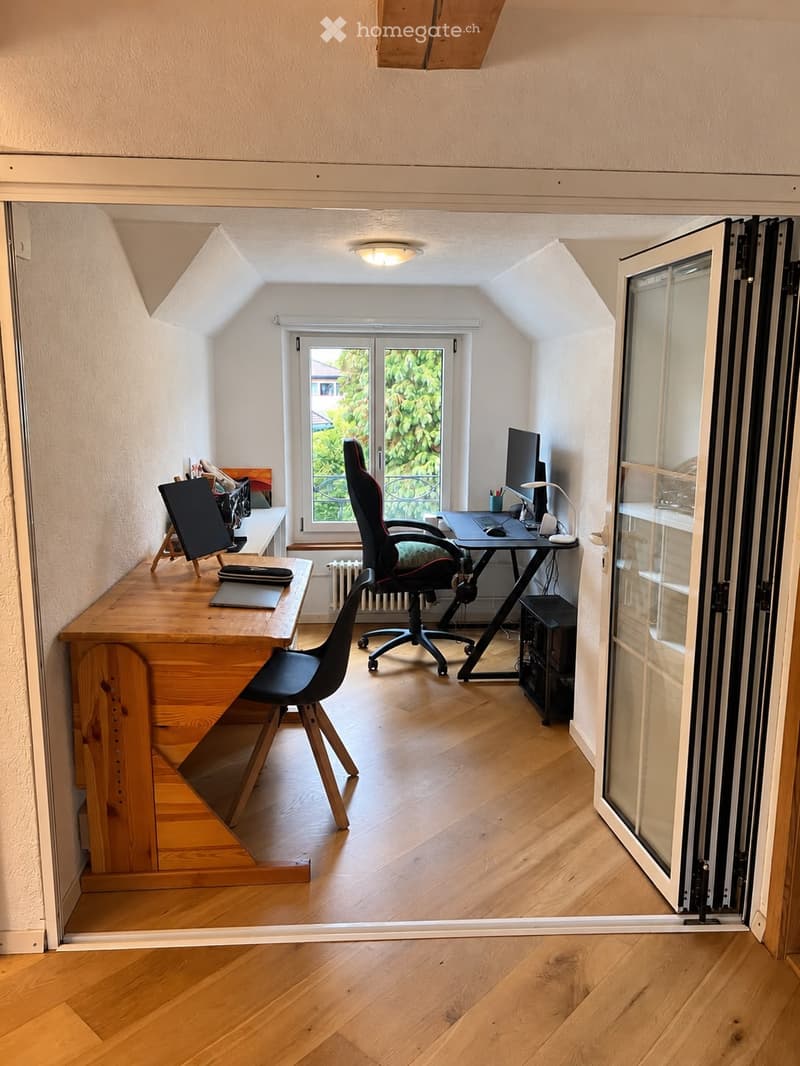 A well-lit, modern workspace with wooden flooring, a large window, a desk with a laptop, a comfortable chair, and a secondary table with a laptop and screen.