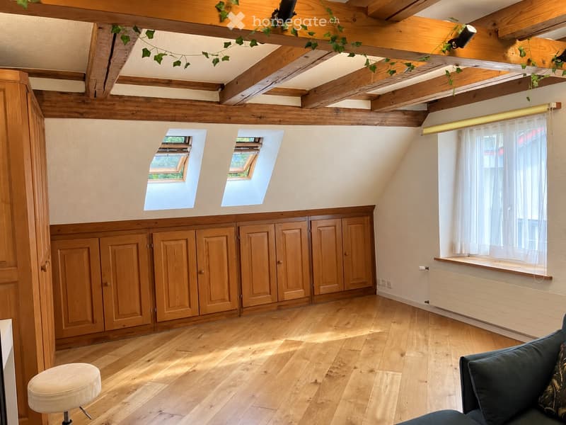 Bright attic room with wooden beams, exposed wooden ceiling, skylights, large window with white curtains, built-in wooden cabinets, and a small sofa.