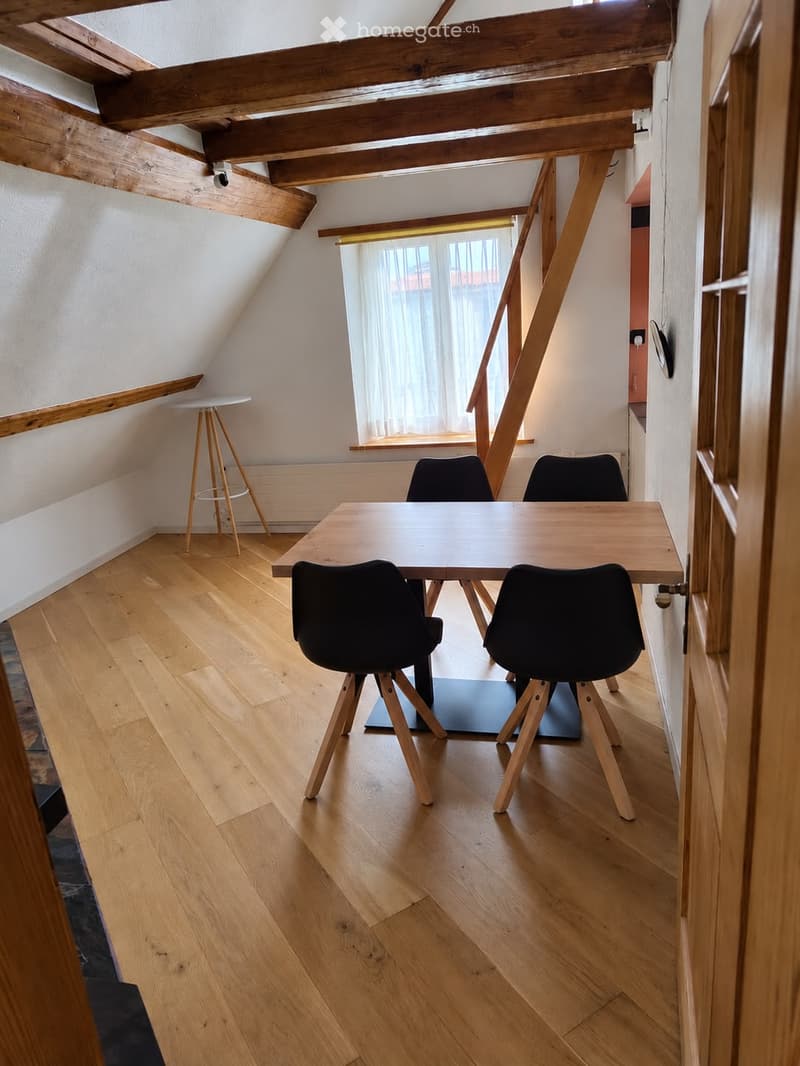Bright, wooden-floored room with slanted wooden ceiling beams, a large window with white curtains, a wooden table with four black chairs, and a small white stool near the window.