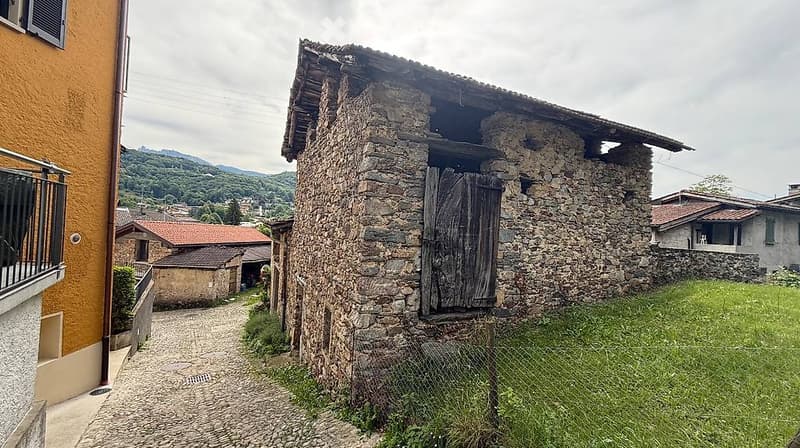 A rustic stone building with a wooden door, situated in a narrow cobblestone street surrounded by other stone houses and greenery.