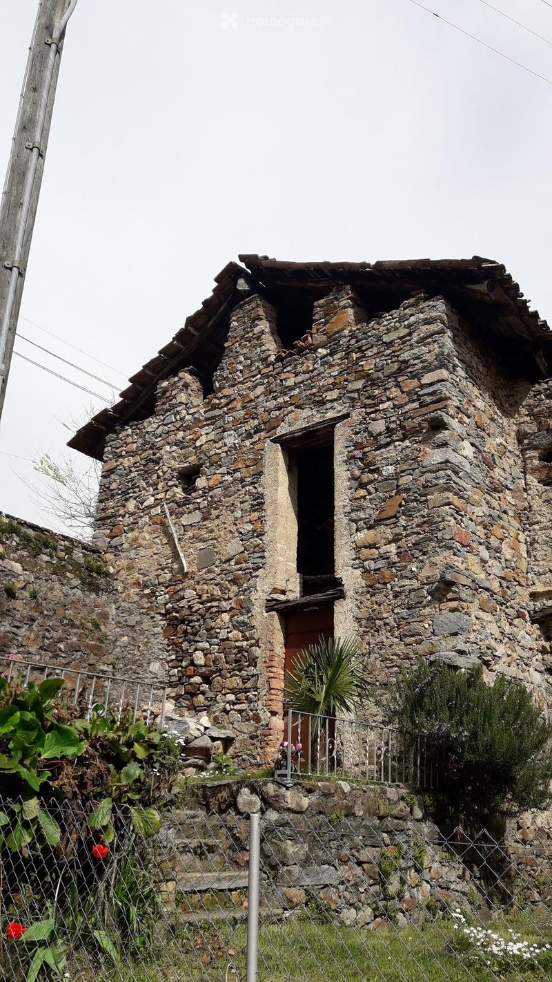 A rustic stone house with a tiled roof, small balcony, and metal railing, surrounded by a stone fence and greenery.