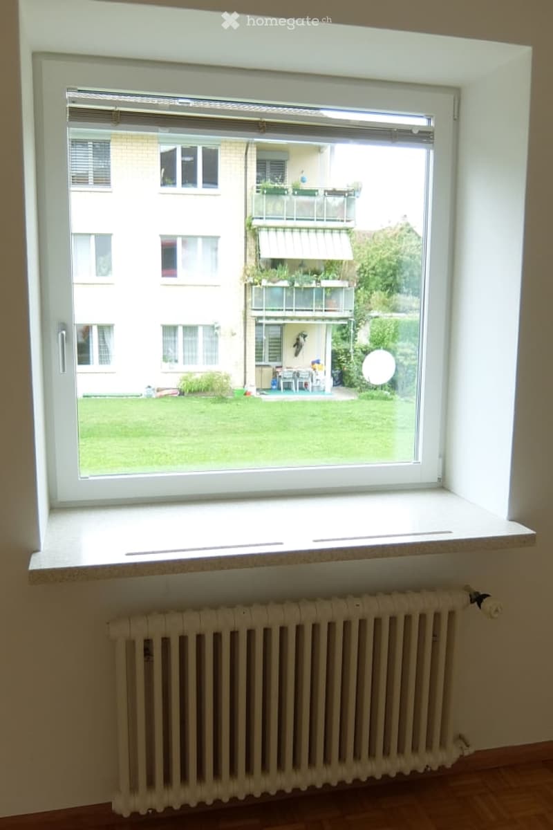 Empty room with wooden flooring, radiator, and large window offering a view of a green lawn, neighboring building, and outdoor furniture.