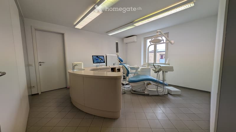 Modern dental office with a reception desk, dental chair, screen, sink, and light fixture.