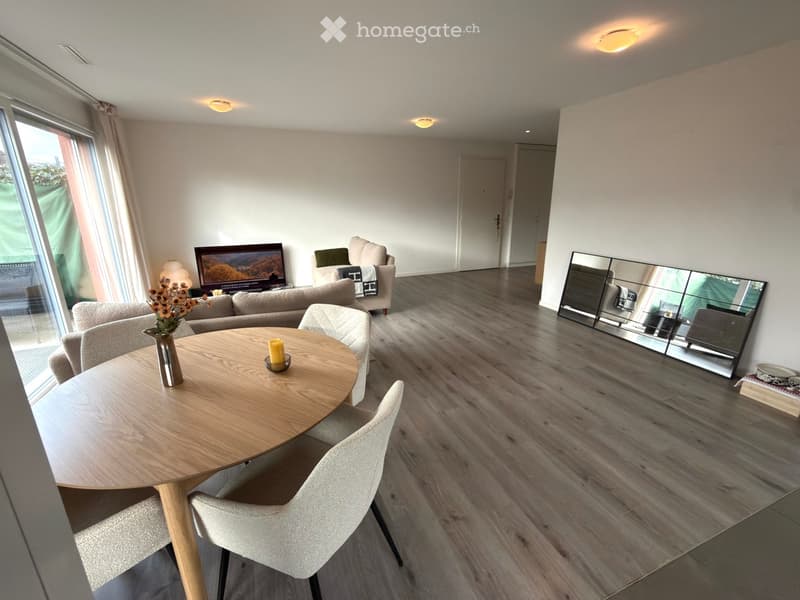 Modern living room with a round wooden table, sofa, chairs, mirror, and television. Features large windows with curtains and a vase with flowers on the table.