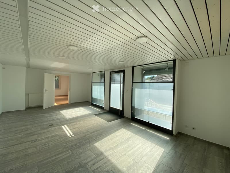Empty room with light-colored walls, wooden floor, and large glass doors leading to an outdoor area. The space is well-lit with recessed ceiling lights.