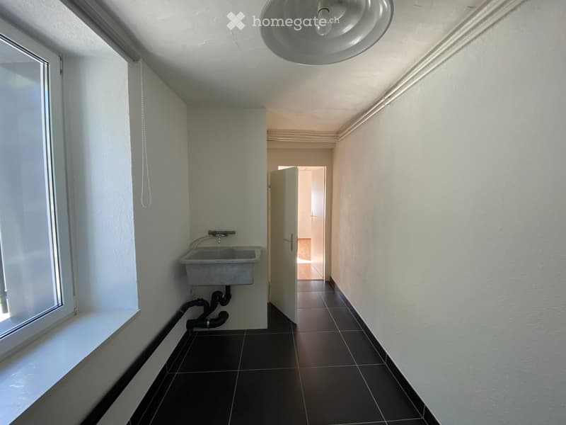 Empty hallway with tiled floor, white walls, a single sink on the left, and a doorway leading to another room.