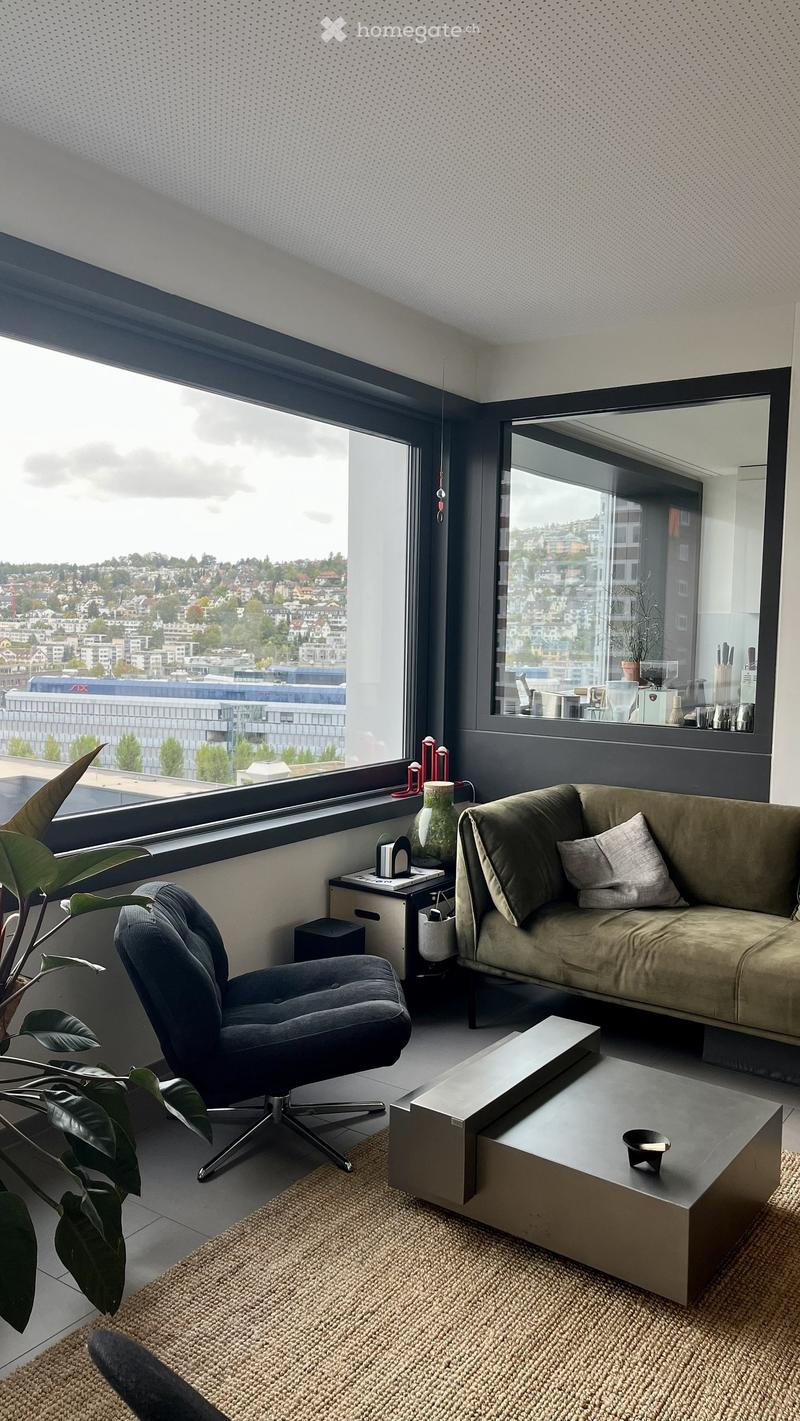 Modern living room with large windows offering a city view, featuring a beige sofa, a dark blue armchair, a rectangular coffee table, a potted plant, and a decorative vase on a side table.