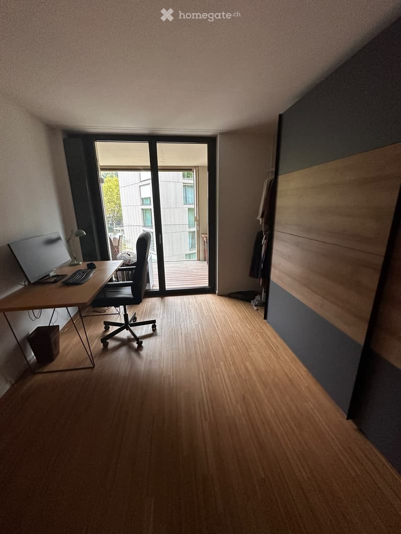 A modern, minimalist office space with wooden flooring, a large window, and a desk setup including a screen, keyboard, mouse, and a chair.