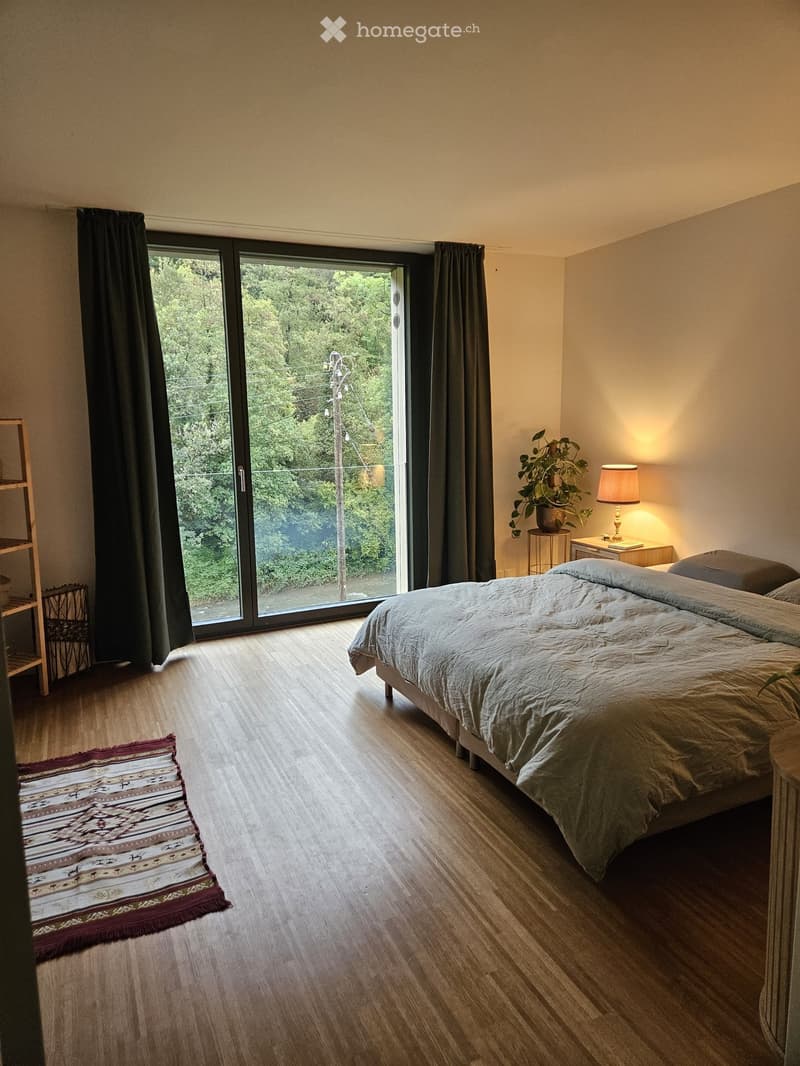 Modern bedroom with wooden flooring, large sliding glass door leading to outdoor view, bed with grey bedding, nightstand with lamp, small plant, and patterned rug.