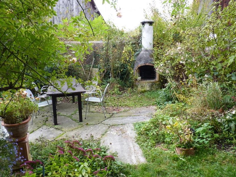 Garden area with a stone pathway, outdoor dining table and chairs, potted plants, and a brick outdoor oven surrounded by lush greenery.